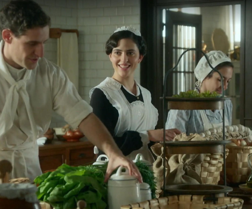 Kucharz Lope i dwie służące, prawdopodobnie Vera, przygotowują posiłki w kuchni pałacu La Promesa, wśród świeżych warzyw i koszy z produktami. Uśmiechnięta kobieta z kokiem spogląda na Lope, podczas gdy druga w tle układa owoce. Więcej o ich losach w hiszpańskim serialu La promesa znajdziesz na Super Seriale.