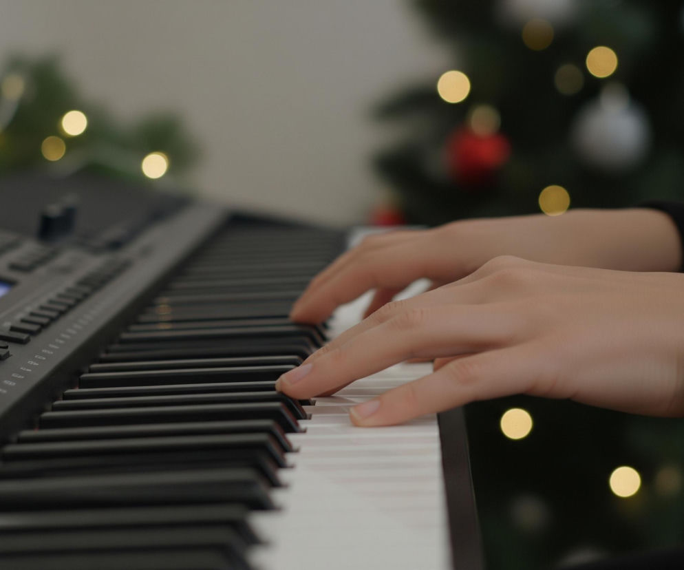 Dłonie w jasnej karnacji grają na klawiaturze elektronicznego instrumentu, składającej się z białych i czarnych klawiszy. Lewa dłoń spoczywa na czarnych klawiszach bliżej panelu sterowania, natomiast prawa dłoń dotyka białych klawiszy, palce są wygięte. Instrument ma czarny panel sterowania z wyświetlaczem i licznymi przyciskami oraz pokrętłami. Tło jest rozmyte, widoczna jest zielona choinka ozdobiona czerwonymi i białymi bombkami oraz ciepłymi, żółtymi światełkami.