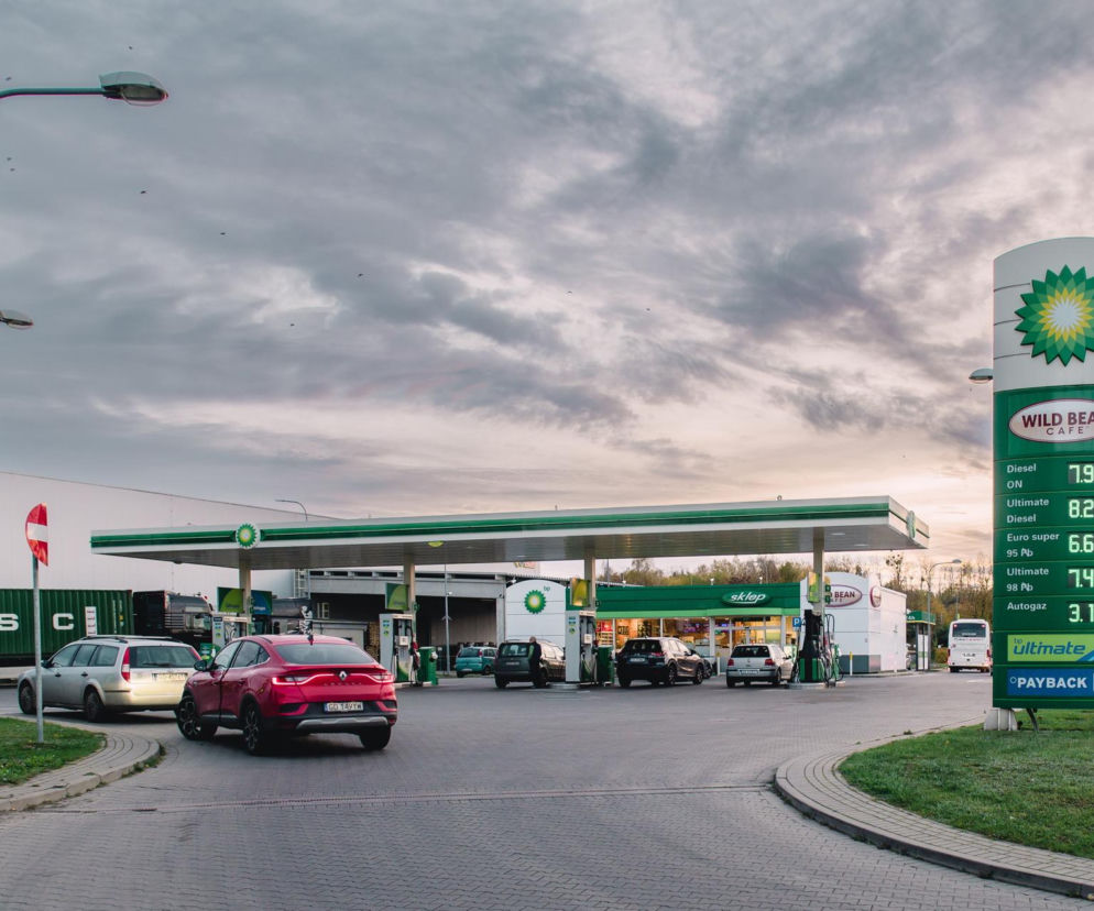 Stacja paliw BP z widocznym pylonem informującym o cenach paliw i zaparkowanymi samochodami, w tym czerwonym Renault. Obraz odzwierciedla kontekst zmian cen paliw, o których można przeczytać na Super Biznes.