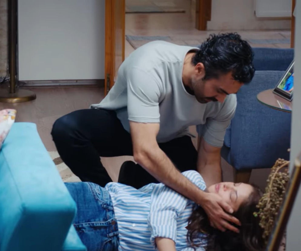 Mężczyzna, Ferit, pochyla się nad nieprzytomną kobietą, Ayse, leżącą na podłodze, z troską podtrzymując jej głowę. Scena dramatyczna z serialu Dziedzictwo, gdzie Ferit ratuje byłą żonę. O emocjonujących odcinkach przeczytasz na Super Seriale.