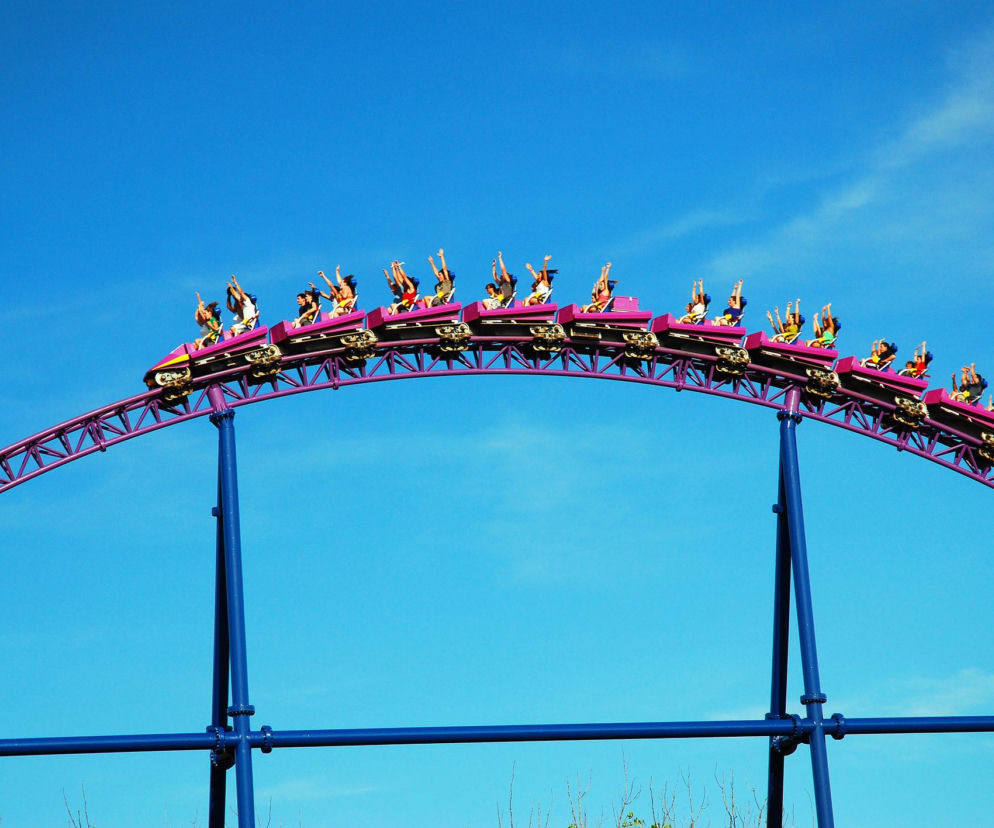 Wagoniki rollercoastera z pasażerami na szczycie fioletowej konstrukcji na tle nieba. O pozwach przeciw parkowi pisze Eska.