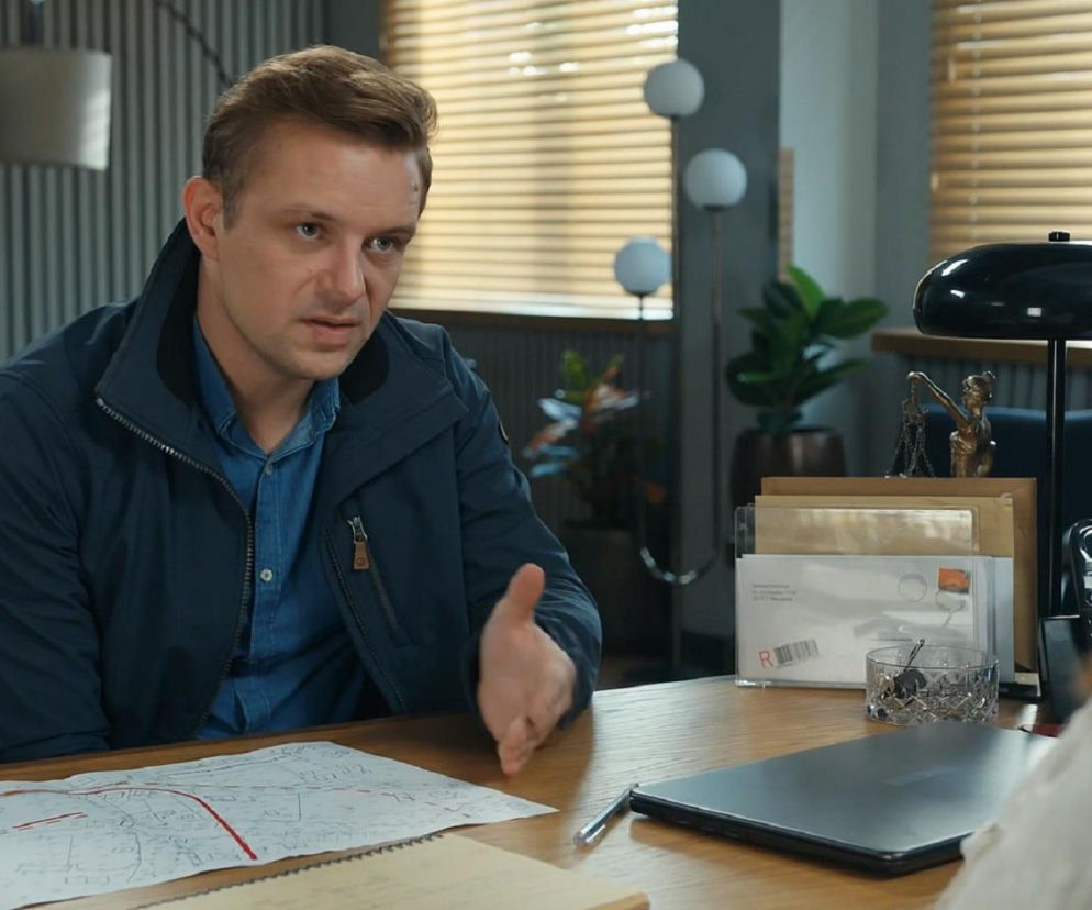 Hubert Pyrka (Marek Molak) z Barw szczęścia siedzi przy biurku, w granatowej kurtce i dżinsowej koszuli, gestykulując ręką, rozmawiając z kimś widocznym od tyłu. Na stole leży mapa i dokumenty. Widok na dalsze losy bohaterów na portalu Super Seriale.