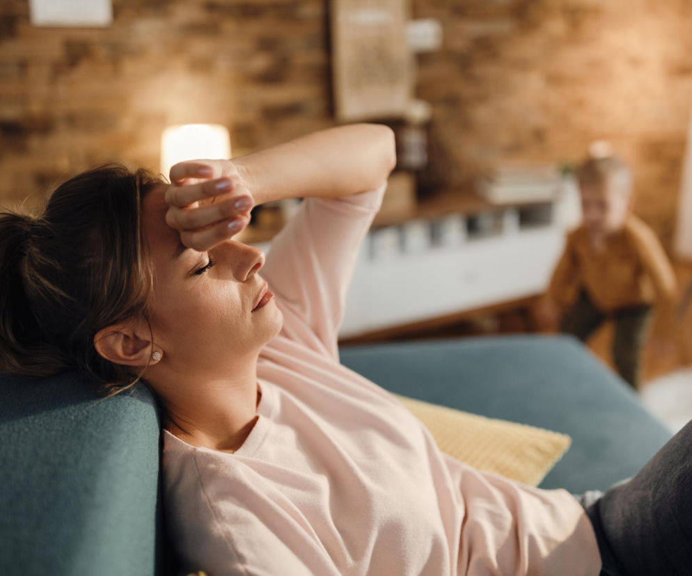 Zmęczona kobieta z ręką na czole leży na kanapie, symbolizując nocne poty i osłabienie. W tle niewyraźne dziecko, co podkreśla trudności w codziennym funkcjonowaniu. O objawach mielofibrozy i innych chorobach przeczytasz na Poradnik Zdrowie.