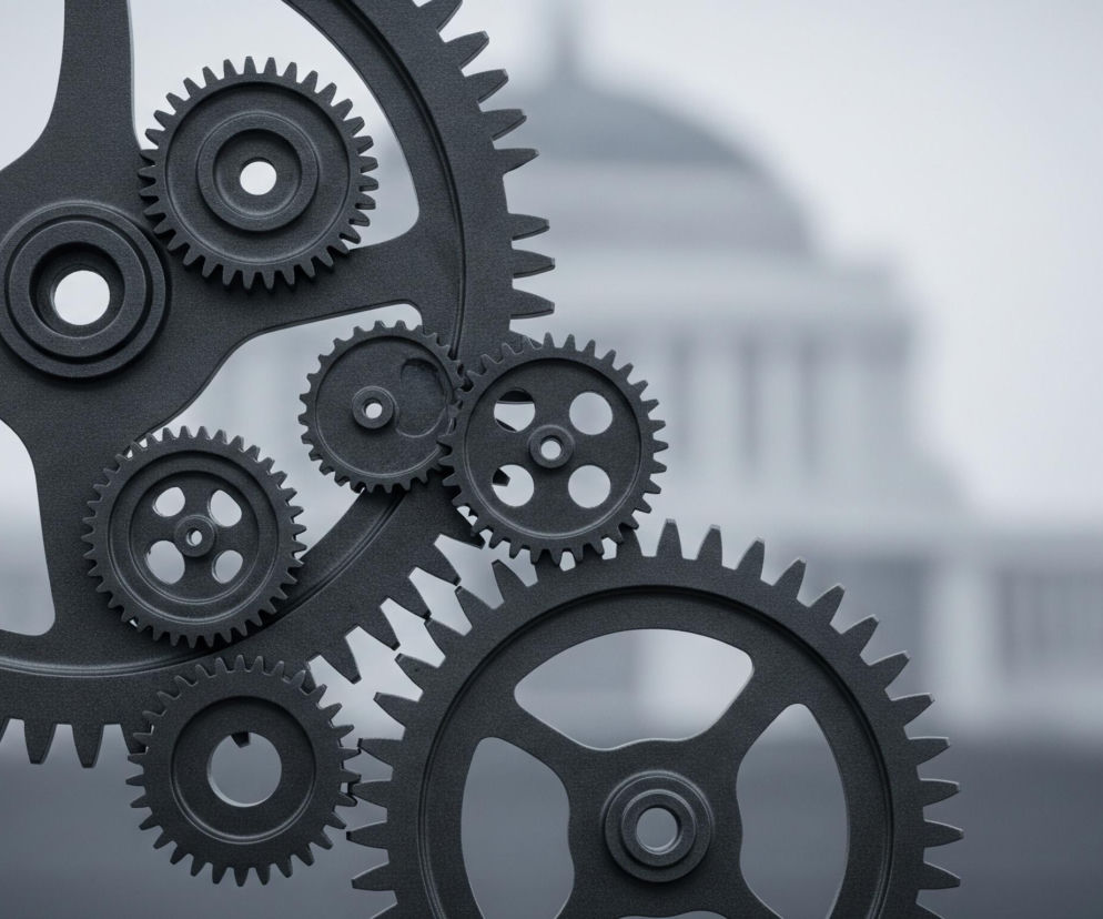 Grupa ciemnoszarych, matowych kół zębatych dominuje pierwszy plan obrazu, tworząc mechaniczny układ. Jedno duże koło z zębami widoczne jest po lewej stronie, z czteroramiennym pająkiem wewnątrz, a w jego górnej części mniejsze koło zębate z centralnym otworem. Po prawej stronie tego dużego koła znajduje się kolejne, nieco mniejsze koło z czterema otworami i zębami, a poniżej niego widać część trzeciego dużego koła zębatego z wyciętymi otworami. W tle, rozmyty i niewyraźny, widać zarys monumentalnego budynku z kopułą, utrzymany w jaśniejszych odcieniach szarości, kontrastujący z ciemniejszymi elementami pierwszego planu.