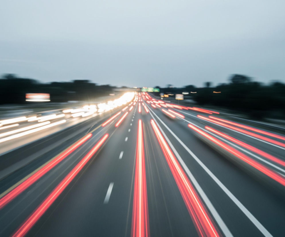 Autostrada z rozmytymi światłami pojazdów zajmuje większość kadru, przedstawiając efekt długiej ekspozycji. Lewa strona drogi wypełniona jest białymi smugami świateł, a prawa strona czerwonymi smugami, symbolizującymi ruch pojazdów w przeciwnych kierunkach. Pasów ruchu jest wiele, oddzielonych białymi, rozmytymi liniami, które zbiegają się w oddali ku horyzontowi. Powyżej drogi widoczne jest rozmyte, szare niebo, a po bokach autostrady ciemne, niewyraźne sylwetki drzew lub budynków.