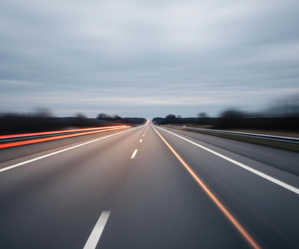 Szeroką autostradę z trzema pasami ruchu w każdym kierunku, oddzielonymi pasem zieleni pośrodku. Droga biegnie prosto w dal, z rozmytym efektem ruchu na poboczach i pasach jezdni, co sugeruje dużą prędkość. Po lewej stronie widoczne są jaskrawoczerwone smugi światła, prawdopodobnie tylne światła mijających pojazdów, natomiast po prawej stronie, na pasie oddzielającym pasy ruchu, widoczna jest pomarańczowa smuga światła, będąca prawdopodobnie reflektorami nadjeżdżających samochodów. Niebo jest pochmurne, w odcieniach szarości i błękitu, z widocznymi poziomymi smugami chmur.
