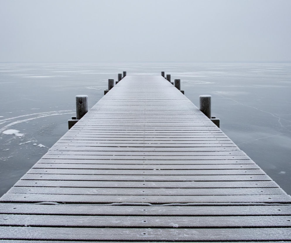 Długi, drewniany pomost pokryty cienką warstwą szronu rozciąga się w głąb zamarzniętej tafli wody, znikając w oddali. Po obu stronach pomostu widać pokryte szronem pale, a powierzchnia lodu jest częściowo pokryta śniegiem i posiada widoczne pęknięcia. Horyzont jest niewyraźny, niebo w kolorze bieli i jasnej szarości łączy się z zamarzniętą powierzchnią.