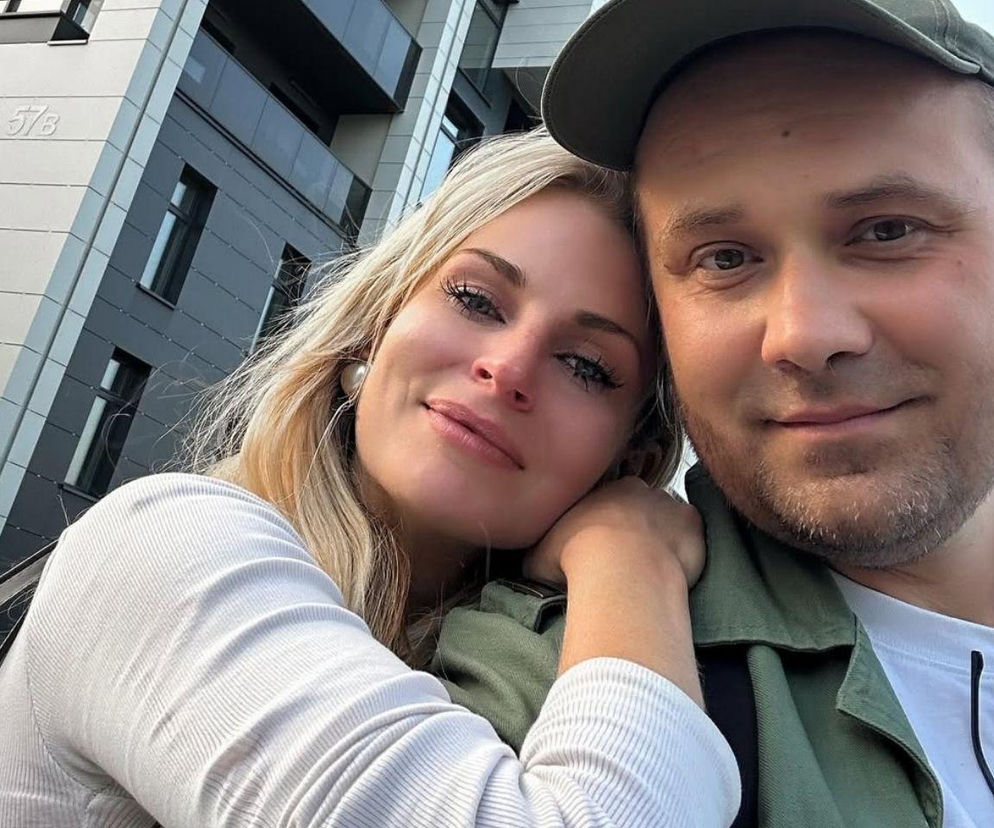 Adrian Szymaniak i jego partnerka w zbliżeniu na tle nowoczesnego budynku, uśmiechają się, prezentując spokój i wsparcie w walce z chorobą. Adrian opowiada o postępach w leczeniu glejaka, co można przeczytać na portalu Poradnik Zdrowie.