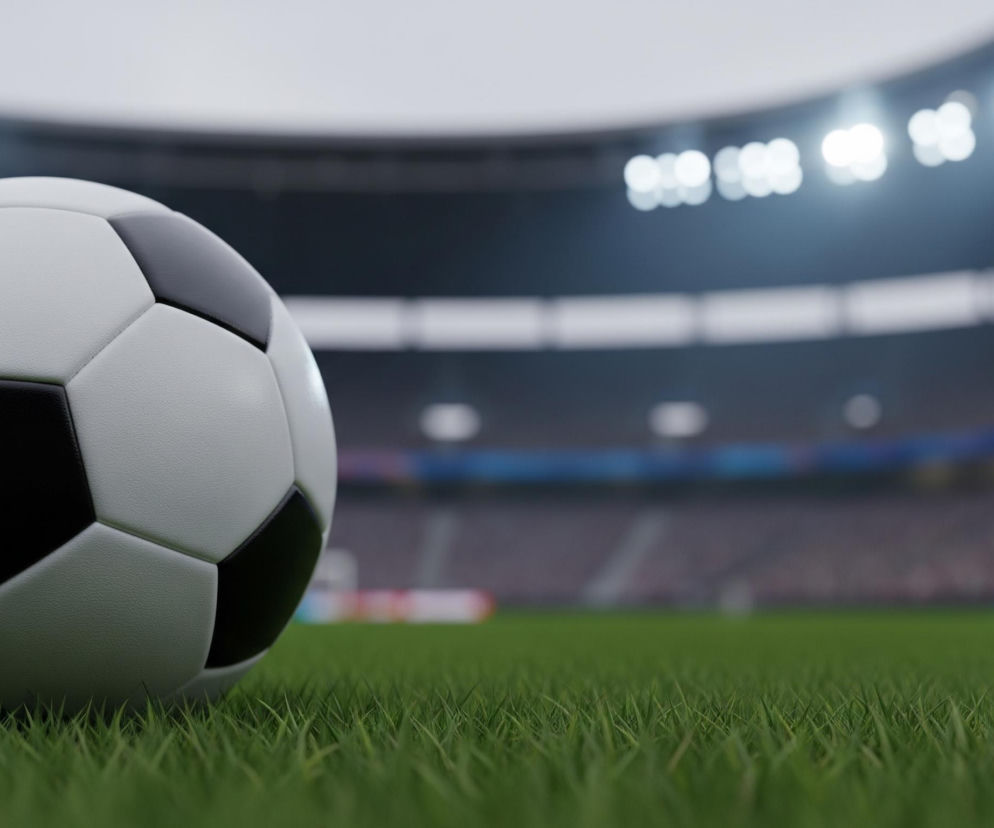 Piłka nożna o tradycyjnym czarno-białym wzorze leży na zielonej trawie na pierwszym planie, jej górna lewa część jest niewidoczna. Futbolówka, składająca się z sześciokątnych i pięciokątnych paneli, jest lekko matowa. W tle, rozmyte, widoczny jest stadion z jaskrawymi światłami reflektorów rozświetlających trybuny, które są ledwie widoczne.