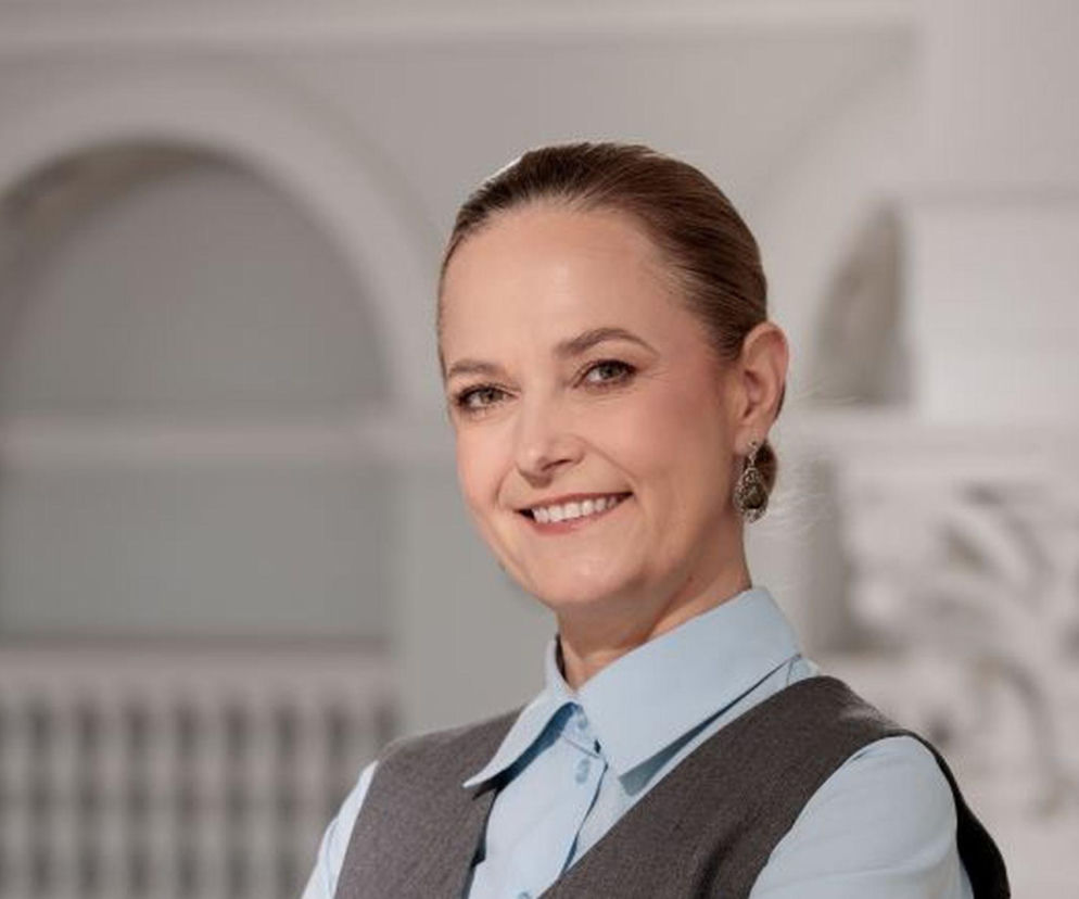 Katarzyna Kęcka, wiceminister zdrowia, uśmiecha się, ubrana w jasnoniebieską koszulę i szarą kamizelkę, na tle architektonicznych detali. O reformach w kształceniu lekarzy przeczytasz więcej na naszym portalu.