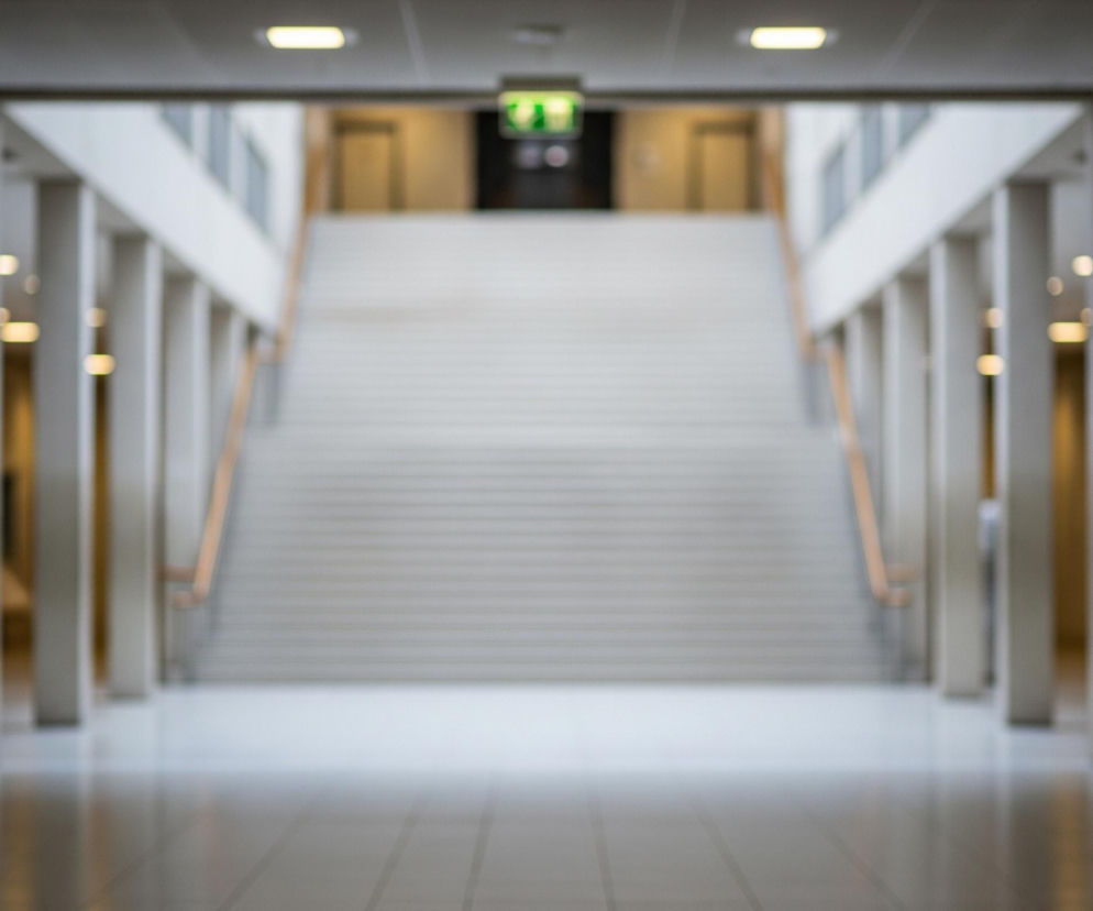 Długi, symetryczny korytarz z rzędem filarów po obu stronach, prowadzący do schodów centralnie umieszczonych na końcu. Schody są szerokie, mają jasne stopnie i drewniane poręcze po obu stronach, a u ich szczytu widoczne są drzwi i zielony znak wyjścia. Całe ujęcie jest nieostre, z centralnym punktem ostrości ustawionym na odległych schodach i znaku wyjścia, natomiast bliższe elementy, takie jak podłoga i filary, są rozmyte. Oświetlenie pochodzi z lamp sufitowych, tworząc refleksy na podłodze, która jest ułożona z jasnych, kwadratowych płytek.