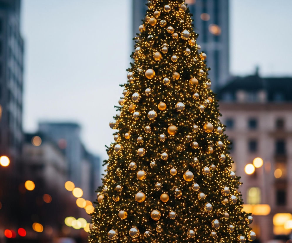 Centralnie ustawiona, dominująca choinka mieści się w kadrze, rozświetlona tysiącami małych, ciepłych, żółtych światełek oraz ozdobiona licznymi bombkami w odcieniach złota i srebra. Drzewko iglaste ma ciemnozielony kolor, a jego kształt jest stożkowaty. Tło stanowią rozmazane, nieostre zabudowania miejskie o szarych i brązowych fasadach, z widocznymi rozproszonymi, okrągłymi światłami w odcieniach żółci, pomarańczy i czerwieni, sugerującymi nocne lub zmierzchowe oświetlenie uliczne.