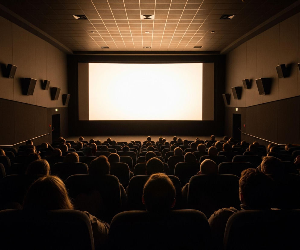 Duża sala kinowa widziana z tyłu, wypełniona publicznością siedzącą w rzędach ciemnych foteli. Jasny, prostokątny ekran dominuje w centrum u góry, rzucając delikatne, ciepłe światło na wnętrze sali i rozświetlając tyły głów oglądających. Boczne ściany, w ciemnym odcieniu brązu, wyposażone są w czarne głośniki, a sufit ma panelową konstrukcję z rzędami kwadratowych kasetonów.