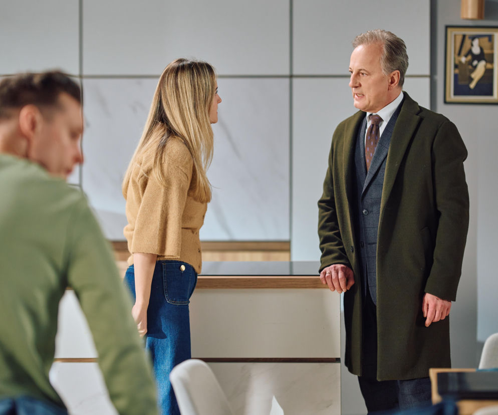 Trzy osoby w nowoczesnej kuchni, w tym mężczyzna w oliwkowym płaszczu i kobieta w brązowym swetrze, z trzecim mężczyzną na pierwszym planie, prawdopodobnie Darek. Scena z serialu Na Wspólnej 4181, gdzie Darek zostaje oskarżony o kradzież z kancelarii, a o tych emocjonujących wydarzeniach możesz przeczytać na Super Seriale.