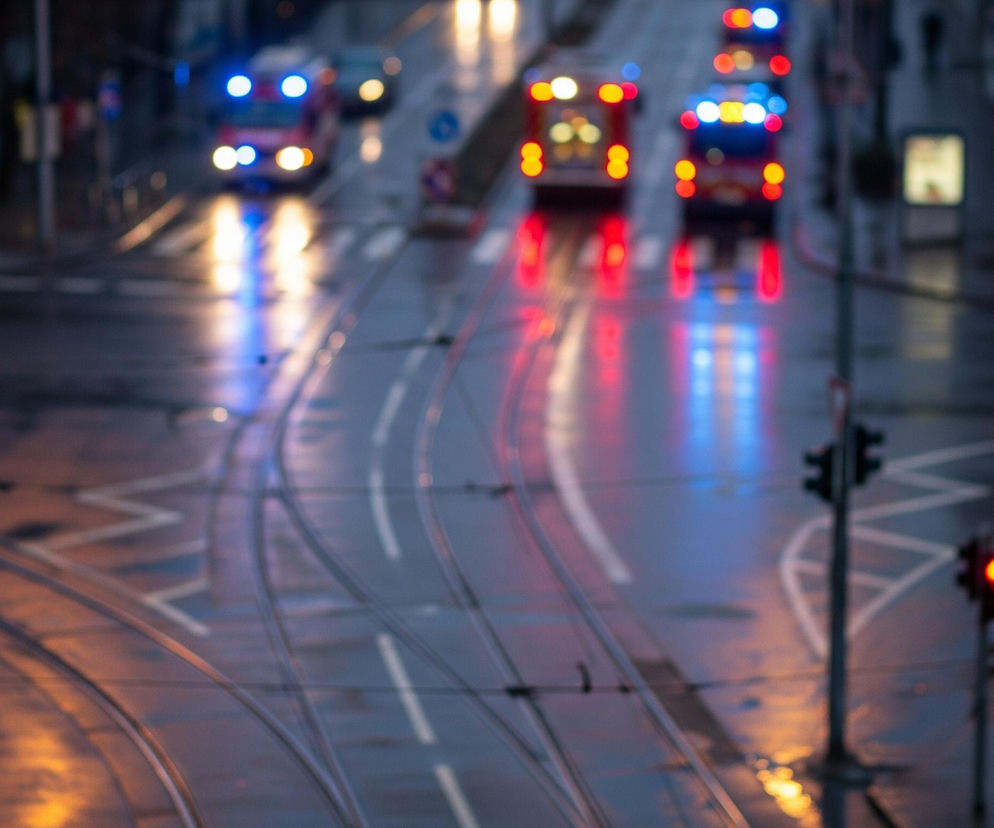 Rozmyty widok z lotu ptaka na miejską ulicę o zmierzchu lub w nocy, z widocznymi torami tramwajowymi i pasami ruchu. Na drodze, w górnej części kadru, znajdują się pojazdy z włączonymi światłami, w tym co najmniej jeden samochód z migającymi niebieskimi światłami sygnalizacyjnymi oraz kilka pojazdów z czerwonymi światłami tylnymi. Światła te tworzą jaskrawe, kolorowe refleksy na mokrej nawierzchni jezdni, dominują czerwone, niebieskie i żółte plamy świetlne. W prawym dolnym rogu widoczny jest słup z włączonym czerwonym światłem sygnalizacji drogowej, a także znaki drogowe w oddali.