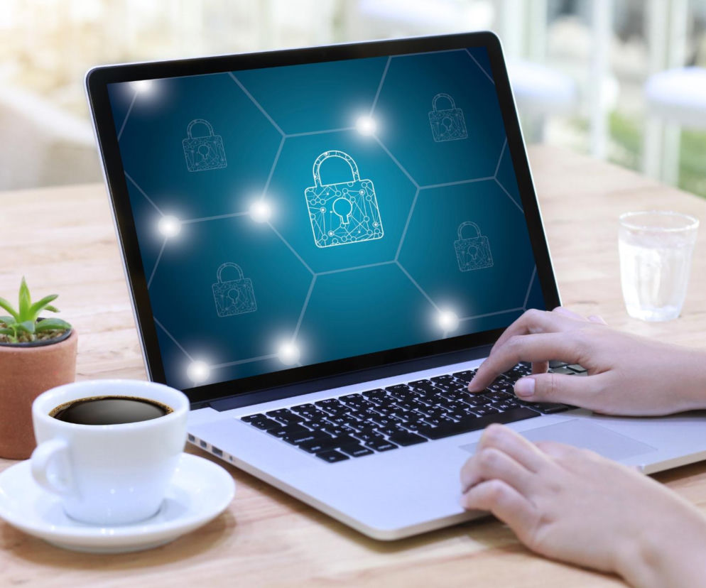 Osoba wpisująca hasło na laptopie, którego ekran pokazuje zabezpieczoną sieć z kłódkami, symbolizującą cyberbezpieczeństwo i ochronę danych. Tło przedstawia kawę i roślinę, artykuły na ten temat znajdziesz na portalu Super Biznes.