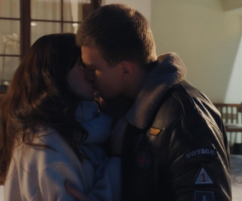 Młodzi Mateusz i Bogna całują się namiętnie na tle domu z oknem, w którym widać inną kobietę – Majkę, stojącą wewnątrz, patrzącą na nich. Mateusz ma na sobie czarną kurtkę z futrzanym kołnierzem, a Bogna jasną bluzę. Ta scena buduje napięcie w M jak miłość i więcej o niej przeczytasz na portalu Super Seriale.