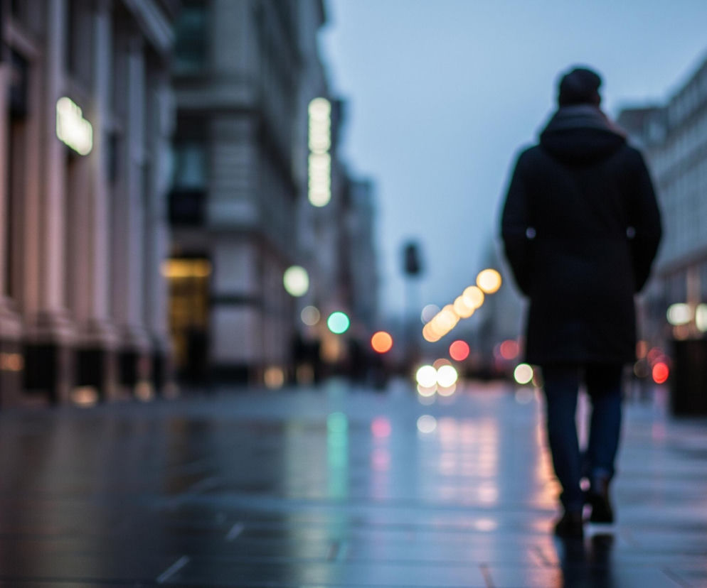 Mężczyzna w ciemnej kurtce z kapturem i czapce idzie ulicą, widoczny od tyłu, jego postać jest nieostra. Ulica jest mokra, odbijając rozmazane światła latarń i sygnalizacji świetlnej w odcieniach pomarańczu, czerwieni, zieleni i bieli. Po lewej stronie widać rozmyte, jasne elewacje budynków z kolumnami, a po prawej stronie wyższe, szare budynki miejskie, również nieostre. Tło to rozmyte, jasnoniebieskie niebo o zmierzchu.