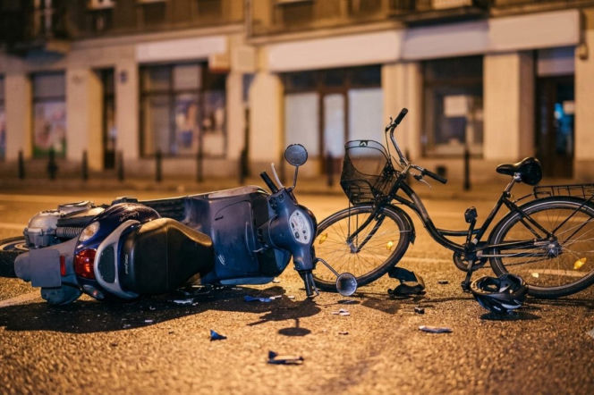 Przewrócony skuter i rower na jezdni w nocy, po wypadku drogowym, na tle niewyraźnych budynków. Widoczne ślady kolizji i fragmenty rozbitych pojazdów, leżący obok kask. Więcej szczegółów o tym zdarzeniu przeczytasz na naszym portalu.