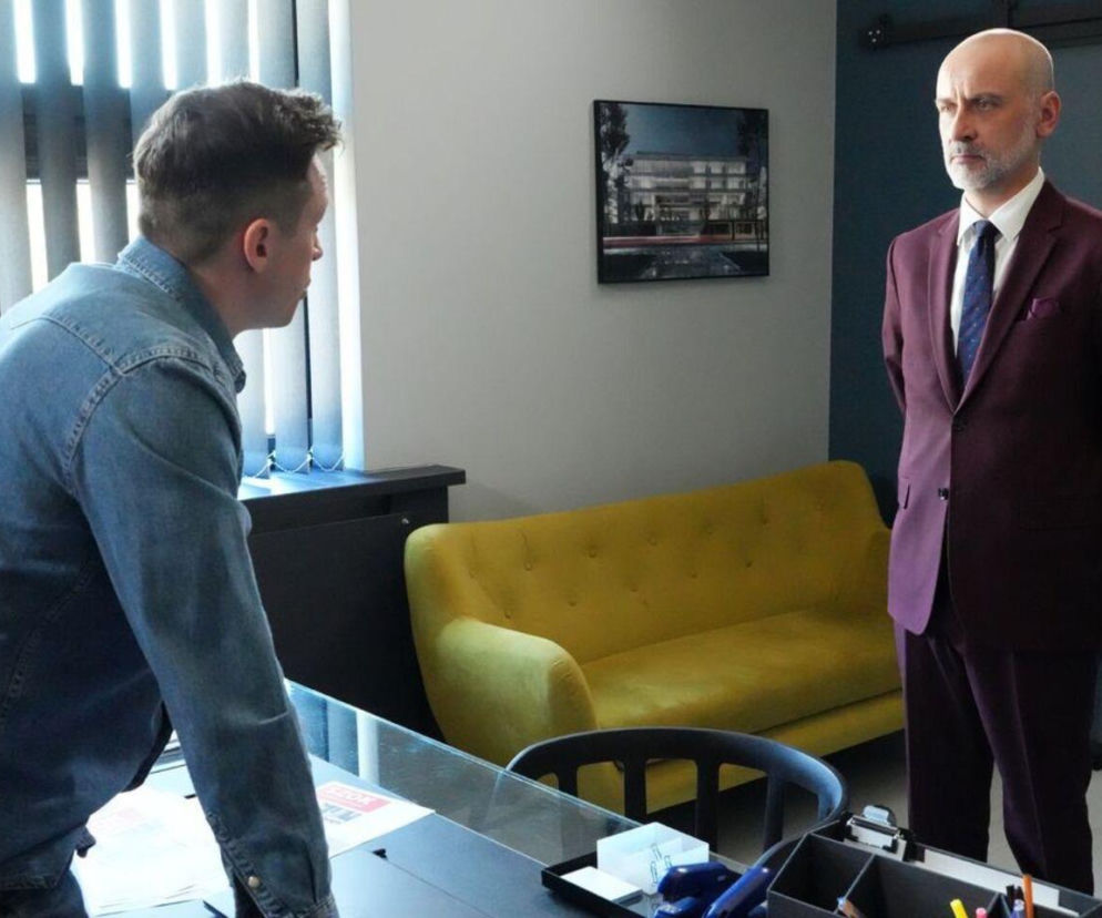 Kornel (Jakub Bohosiewicz) w bordowym garniturze stoi naprzeciw Modrzyckiego (Piotr Ligineza) w jeansowej koszuli, pochylonego nad biurkiem. Widoczny w tle żółty fotel i regał. Więcej o ich losach na Super Seriale.