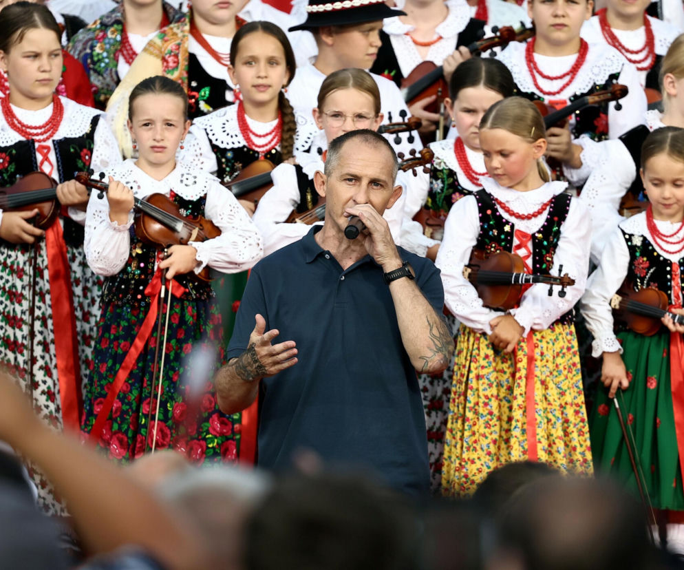Raper Eldo śpiewa do mikrofonu na tle dzieci w strojach ludowych. O jego występie dla prezydenta przeczytasz w Eska.