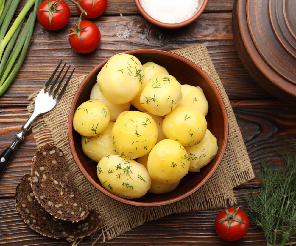 Gotowane młode ziemniaki z koperkiem w misce, otoczone świeżymi warzywami: pomidorami, szczypiorkiem i koperkiem, z solniczką i pieczywem na drewnianym tle, ilustrujące artykuł Poradnik Zdrowie o soleniu ziemniaków przy nadciśnieniu.