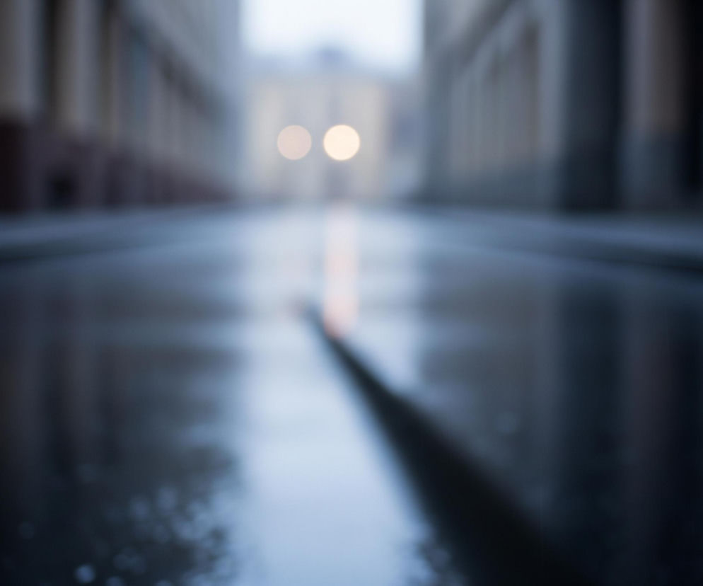 Głębokie rozmycie obrazu przedstawia scenę miejską o zmierzchu lub wczesnym świcie. W centrum kadru, na horyzoncie, widoczne są dwa jasne, rozmazane punkty świetlne, prawdopodobnie reflekt samochodu, od których biegnie w dół, przez środek kadru, pomarańczowa smuga odbicia na mokrej nawierzchni. Po obu stronach smugi, nawierzchnia ulicy jest ciemna, mokra, z delikatnymi jaśniejszymi odbiciami. Po bokach, poza ostrością, widoczne są zarysy budynków, z dominującymi pionowymi formami i poziomymi liniami, sugerującymi fasady. Cała kompozycja utrzymana jest w chłodnych odcieniach szarości i błękitu, z akcentami pomarańczowego światła.