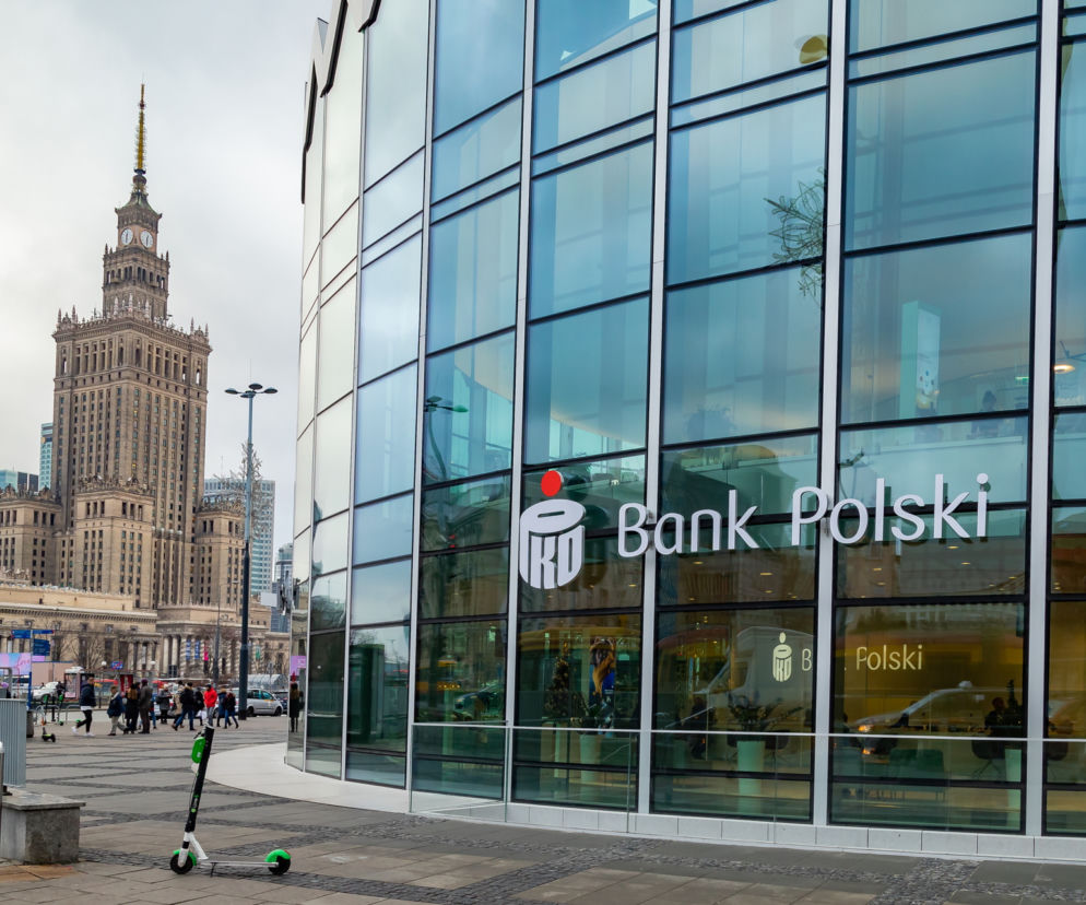 Fasada PKO Banku Polskiego z logo na tle nieba, po lewej stronie Pałac Kultury i Nauki w Warszawie, a na pierwszym planie hulajnoga. Zdjęcie ilustruje temat opóźnień przelewów w święta, o czym przeczytasz na Super Biznes.