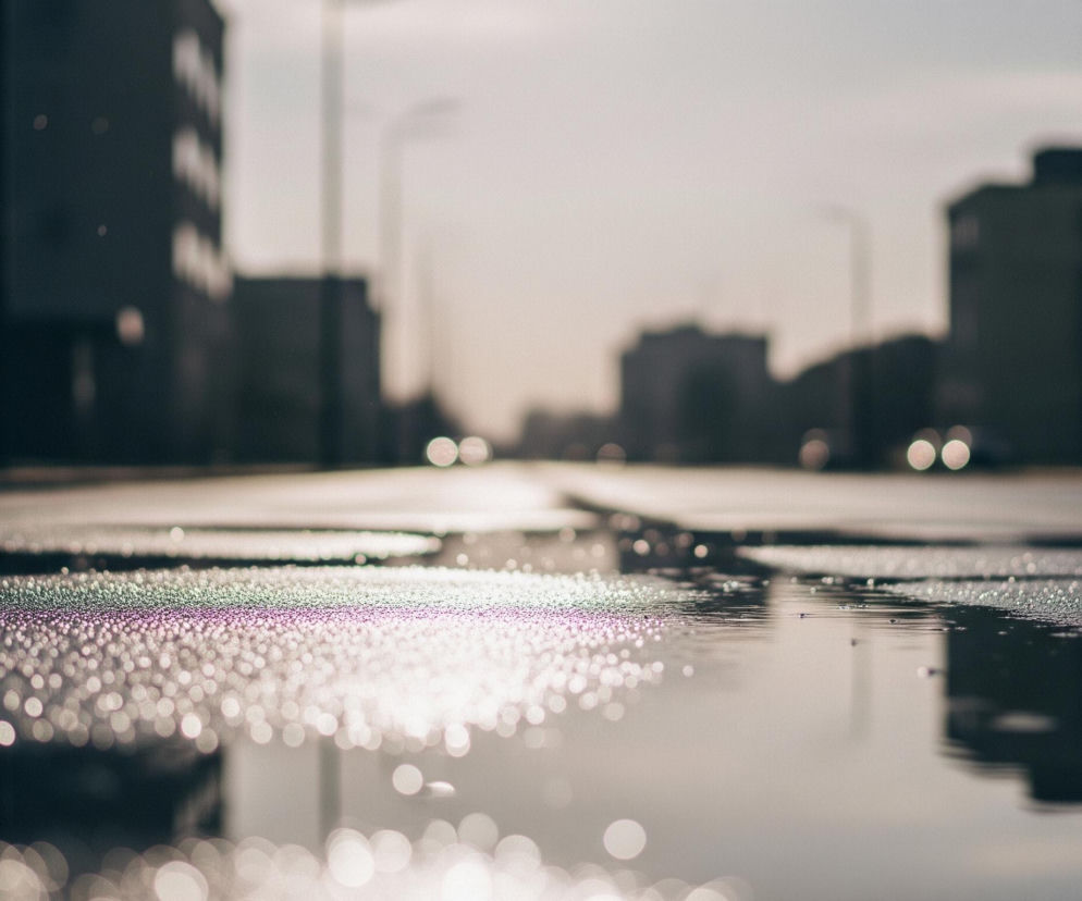 Na pierwszym planie widoczna jest mokra powierzchnia asfaltu z licznymi, błyszczącymi kroplami wody, które tworzą efekt bokeh. W centrum kadru, na powierzchni kałuży, odbija się rozmyte światło, tworząc jasną plamę. Dalej w tle rozciąga się miejska ulica z rozmazanymi sylwetkami budynków po obu stronach i odległymi światłami samochodów lub latarni. Cały obraz utrzymany jest w stonowanych, szaro-brązowych barwach, z delikatnymi fioletowymi refleksami w kroplach wody.
