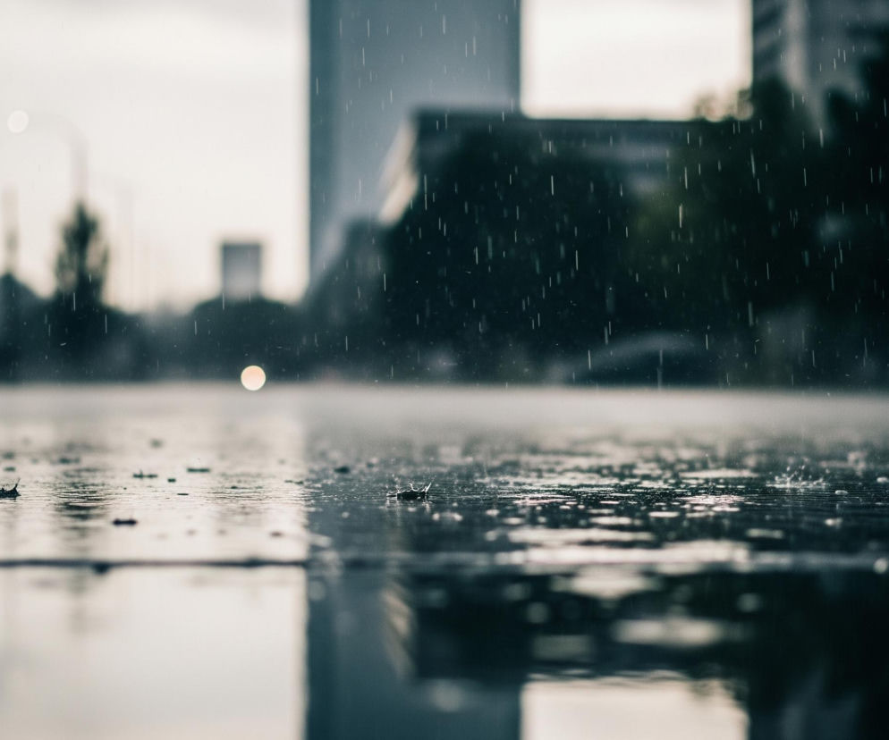 Głównym obiektem jest mokra, ciemna nawierzchnia z odbijającymi się w niej światłami. Na pierwszym planie widać krople deszczu uderzające o powierzchnię, tworząc drobne rozbryzgi i fale. Po lewej stronie leży suchy liść. W tle, rozmyte i niewyraźne, rysują się kontury miejskich budynków i drzew. Jasna, okrągła plama światła, prawdopodobnie latarni, świeci w centralnej części obrazu, a po lewej stronie widoczne jest czerwone światło. Całość utrzymana jest w chłodnych, szaro-niebieskich odcieniach, z pionowymi smugami deszczu widocznymi w powietrzu.