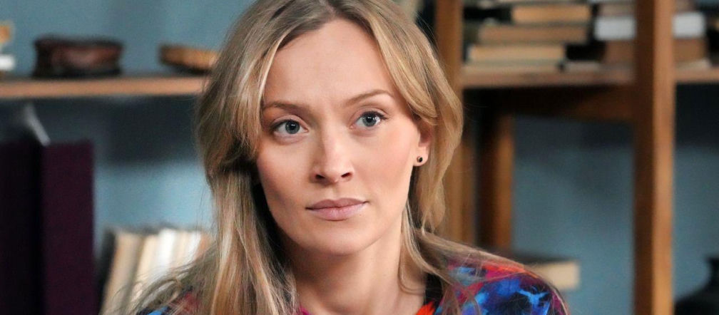 Portret kobiety o blond włosach, Renaty (Anna Mrozowska) z serialu „Barwy szczęścia”, z tłem półek z książkami. Ma na sobie kolorową bluzkę, a jej twarz wyraża zamyślenie, co idealnie oddaje emocje związane z jej powrotem do pracy. Więcej o losach Renaty przeczytasz na Super Seriale.