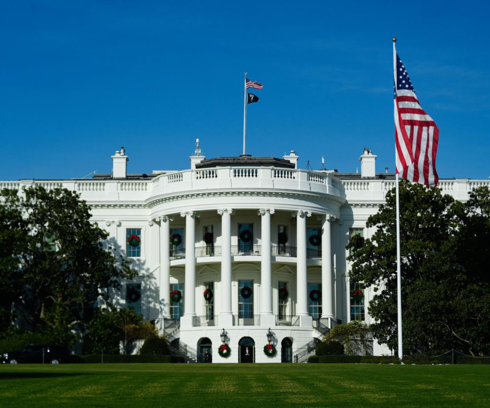 Biały Dom udekorowany świątecznymi wieńcami na tle błękitnego nieba. Widać powiewającą flagę USA. Artykuł na Architektura Murator Plus opisuje kontrowersje wokół zmian w Białym Domu, w tym nowej sali balowej.