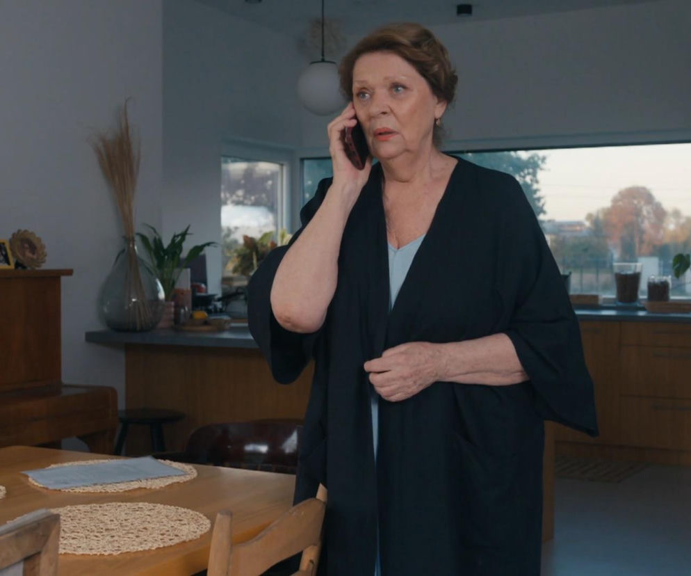 Józefina (Elżbieta Jarosik) z serialu Barwy szczęścia w domowym wnętrzu, ubrana w czarną narzutkę i jasną bluzkę, prowadzi rozmowę telefoniczną z zaniepokojoną miną. Jej rozpaczliwy telefon do Cezarego, o którym przeczytasz na portalu Super Seriale, ma na celu ratowanie rozpadającego się małżeństwa syna.