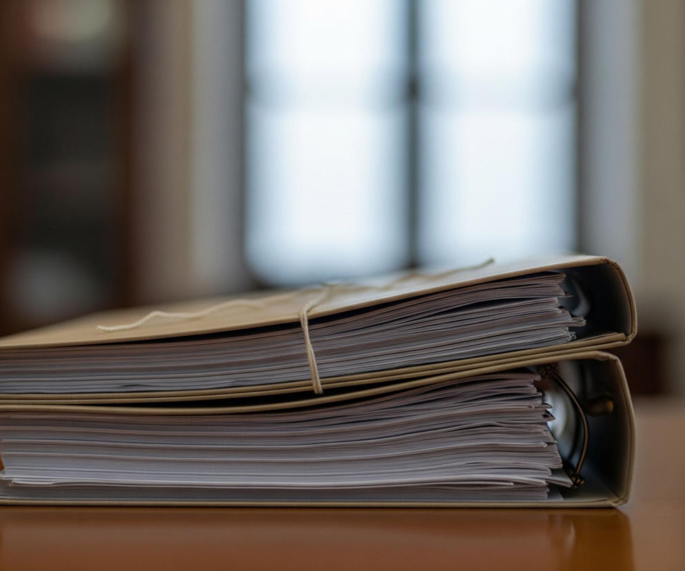 Dwa stosy dokumentów spiętych w teczki leżą na jasnobrązowym, drewnianym biurku. Górna teczka, w kolorze beżowym, jest przewiązana jasnym sznurkiem, a jej wnętrze wypełniają gęsto ułożone białe kartki papieru. Pod nią znajduje się kolejna, podobna teczka z widocznym mechanizmem ringowym. Tło jest rozmyte, z ciemnymi obszarami po lewej stronie i jasnym, nieostrym oknem po prawej.
