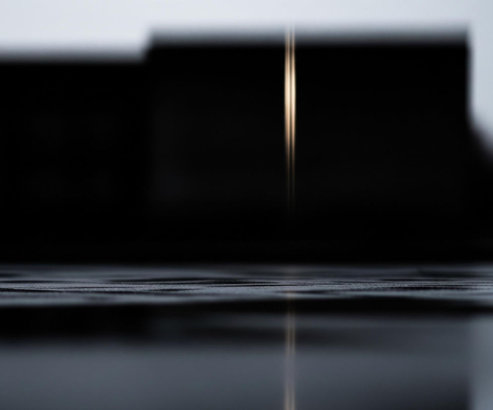 Na pierwszym planie rozciąga się ciemnoszara, wilgotna powierzchnia z widocznymi poziomymi, równoległymi liniami tekstury, a jej dolna część silnie odbija światło, tworząc rozmyte lustrzane odbicie. W tle, na wysokości horyzontu, dominuje ciemna, zamazana sylwetka dwóch prostokątnych obiektów, z jasnym pionowym pasem światła odbijającym się pomiędzy nimi. Po prawej stronie, na wysokości tła, majaczy rozmyta, czerwona plama światła, która również odbija się w dolnej, lustrzanej części obrazu. Całość utrzymana jest w chłodnych, ciemnych barwach z dominacją czerni, szarości i bieli, przełamanych jedynie złotym i czerwonym akcentem świetlnym.