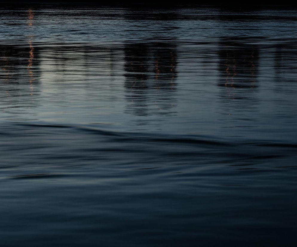 Większość kadru zajmuje ciemna, spokojna woda o granatowo-szarych odcieniach, z delikatnymi falami i zmarszczkami na powierzchni, które rozmywają się ku dołowi. Na górnej części obrazu, na wodzie, widoczne są rozmyte, pionowe odbicia jaśniejszych, pomarańczowych świateł oraz ciemnych, abstrakcyjnych kształtów, sugerujących odbicie odległych obiektów. Kolory są głównie chłodne i stonowane, z akcentami ciepłych pomarańczowych refleksów.