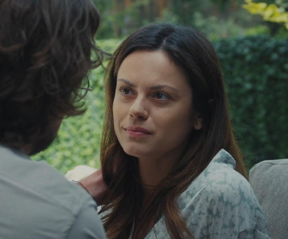 Aktorka Paulina Lasota jako Kasia z serialu M jak miłość w jasnej koszuli w kwiatowe wzory, rozmawia z Mariuszem w ogrodzie. Kasia ma poważny wyraz twarzy. Scena oddaje napiętą atmosferę w związku. Więcej o ich losach na Super Seriale.