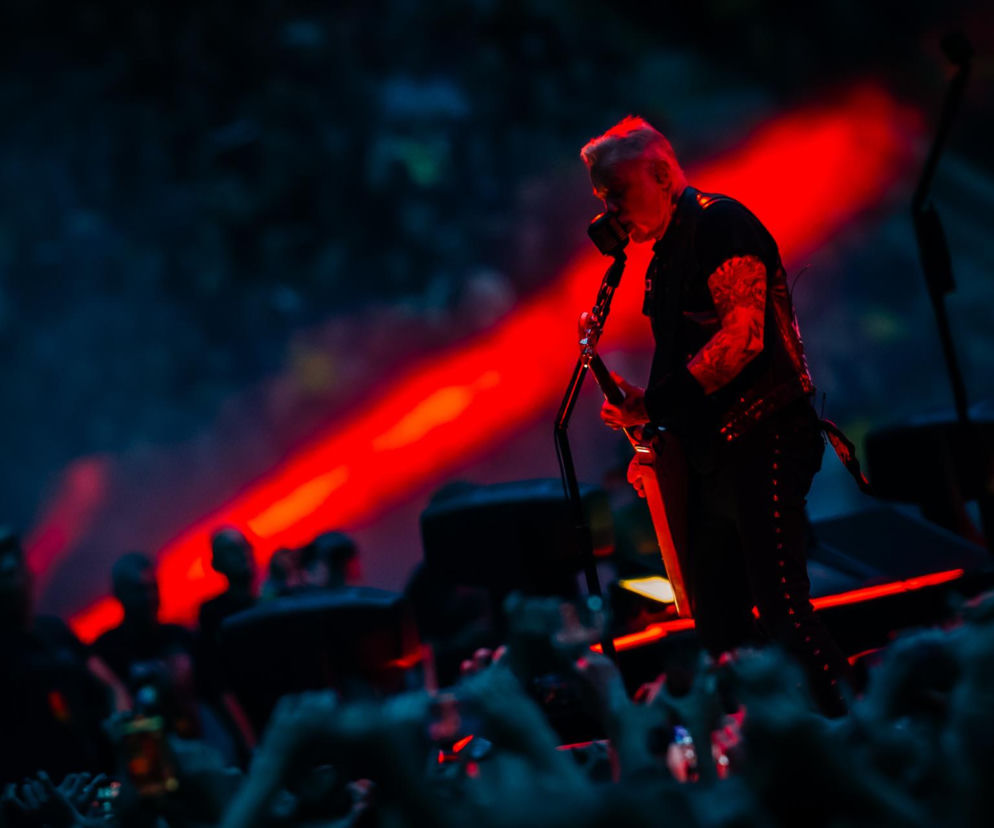James Hetfield z Metalliki śpiewa do mikrofonu na ciemnej scenie. O trasie M72 World Tour przeczytasz na EskaRock.