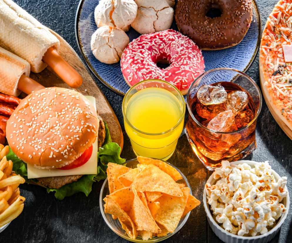 Stół zastawiony niezdrowymi produktami: hamburgerem z frytkami, hot dogami, pizzą, pączkami, ciasteczkami bezowymi, nachos, popcornem, sokiem i napojem gazowanym, które mogą osłabiać leki na nadciśnienie. Sprawdź na Poradnik Zdrowie, czego unikać w diecie.