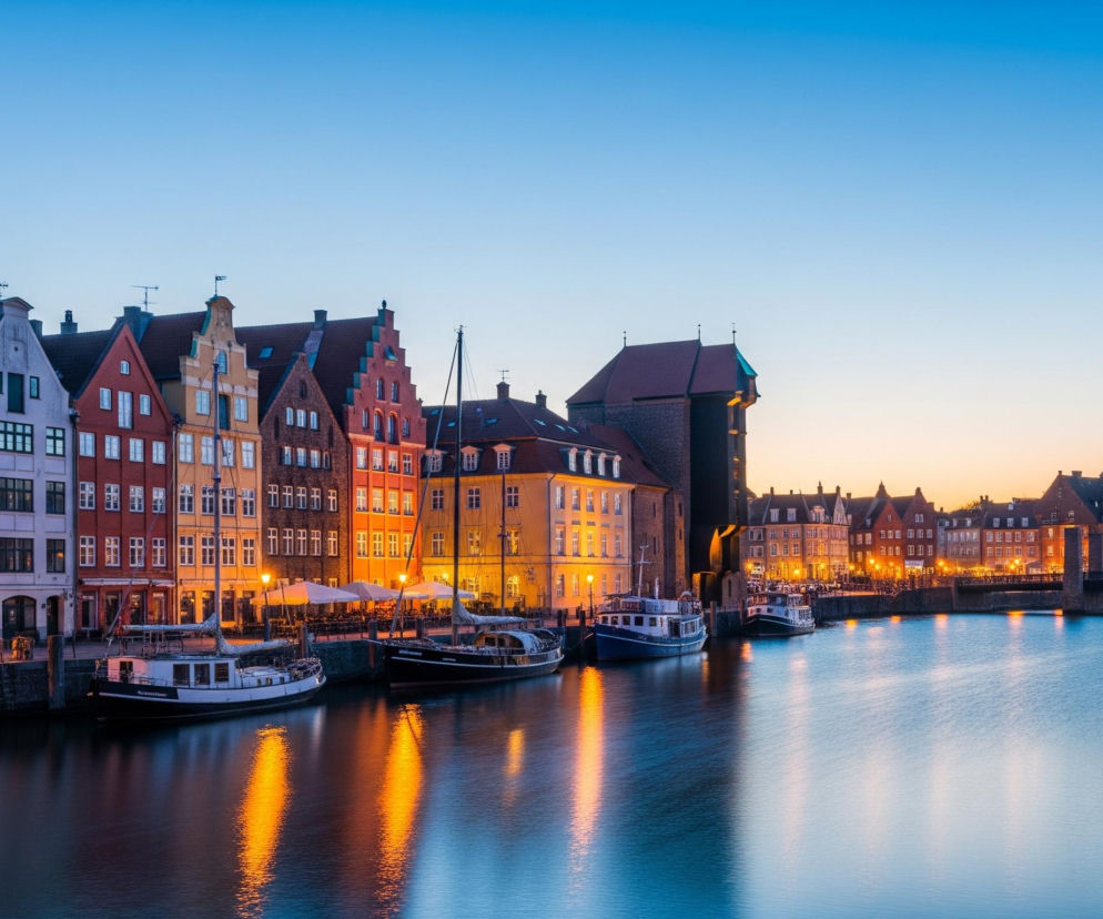 Długi, spokojny kanał lub rzeka przepływa przez centrum kadru, odbijając światła budynków i latarni ulicznych w złotych smugach na powierzchni wody. Wzdłuż lewego brzegu kanału rozciąga się rząd kolorowych, tradycyjnych kamienic o spadzistych dachach, w różnych odcieniach czerwieni, pomarańczy, żółci i bieli, z oświetlonymi oknami i restauracjami na parterach. Przy nabrzeżu zacumowane są różne łodzie i jachty, a w tle widać ciemną konstrukcję dźwigu portowego oraz dalsze budynki. Całość znajduje się pod błękitno-pomarańczowym niebem o zmierzchu, gdzie błękit przechodzi w jaśniejszy odcień horyzontu.