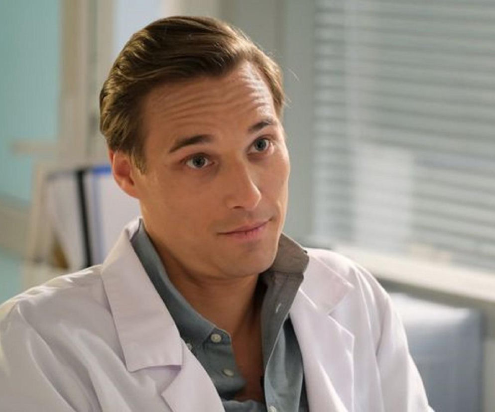 Aktor Mikołaj Krawczyk w roli Sowińskiego, antagonisty w serialu M jak miłość. Sowiński, nowy szef Olka, patrzy z ironicznym uśmiechem, ubrany w niebieską koszulę i biały kitel. Tło stanowią pionowe żaluzje. Więcej o wątku w Super Seriale.