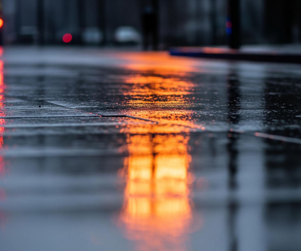 Ulica pokryta deszczem odbija intensywne, rozmyte światła w ciemnym otoczeniu. Na lewo, odblaski czerwonych i niebieskich świateł, pochodzące prawdopodobnie z pojazdu uprzywilejowanego, tworzą pionowe pasy na mokrej nawierzchni. Po prawej stronie, pomarańczowe światło rozlewa się szeroką smugą, kontrastując z chłodnymi tonami otoczenia. Droga na pierwszym planie jest wyraźnie widoczna, pokryta drobnymi kroplami wody, z horyzontem zamazanym w tle.