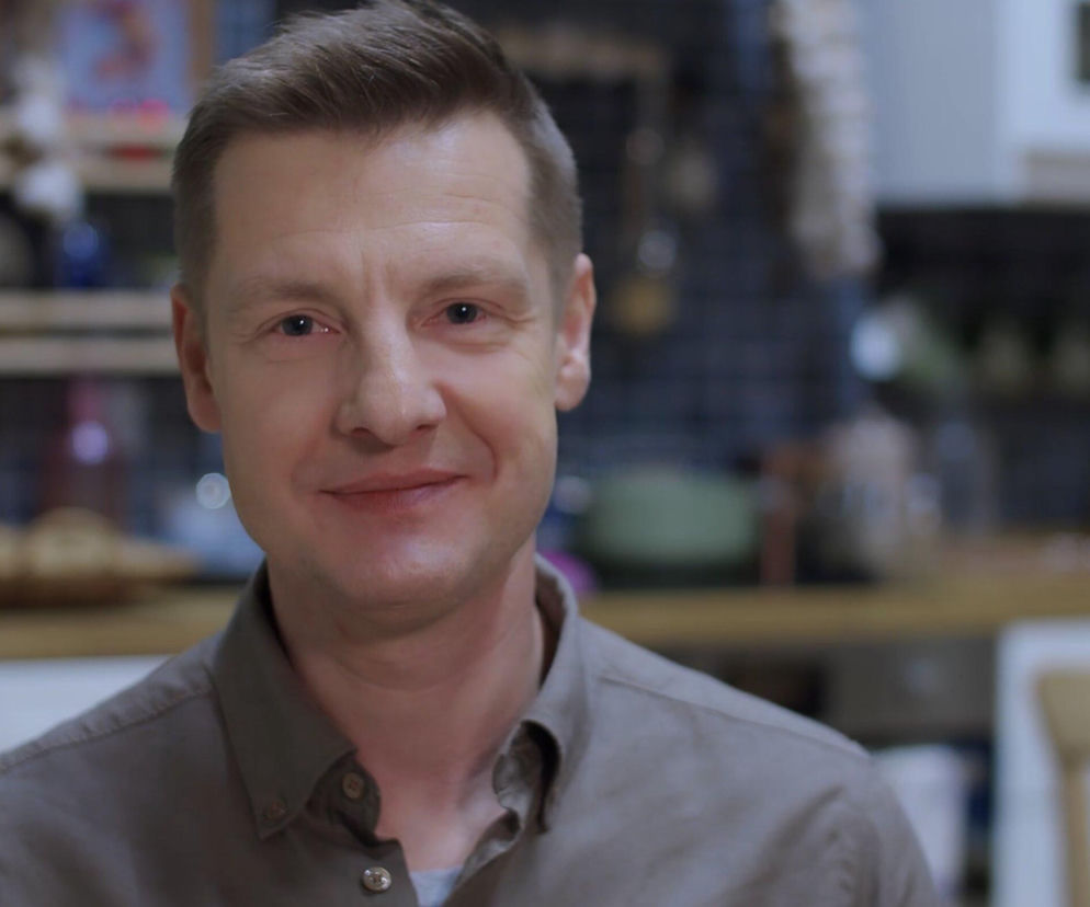 Uśmiechnięty Rafał Mroczek w zbliżeniu na twarz, ubrany w szarą koszulę, na tle rozmytej kuchni. Aktor promuje nowe wątki postaci Pawła Zduńskiego w M jak miłość. Więcej o losach bohatera przeczytasz na portalu Super Seriale.