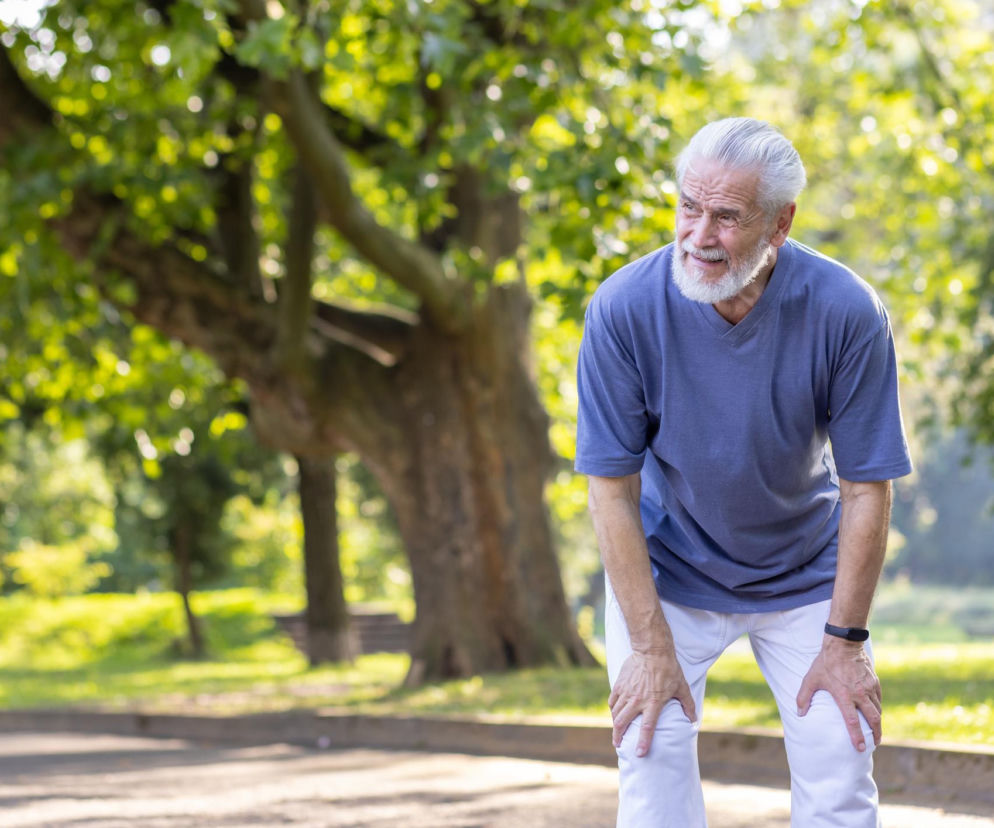 Starszy mężczyzna z siwą brodą i włosami, w niebieskiej koszulce i białych spodniach, stoi pochylony z rękami opartymi na kolanach, zmęczony po wysiłku fizycznym w parku. Wyraźne objawy duszności i wyczerpania, które mogą świadczyć o rzadkiej chorobie serca. Więcej informacji na ten temat znajdziesz na Poradnik Zdrowie.