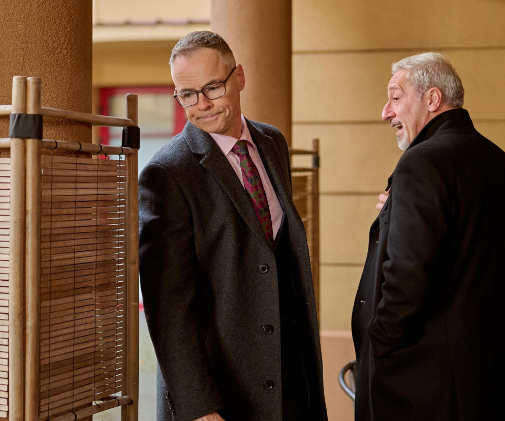 Damian Cieślik w ciemnym płaszczu i okularach z miną zażenowania patrzy na Krupskiego, który z otwartymi ustami, zaskoczony, wpatruje się w przestrzeń obok siebie. Scena z serialu Na Wspólnej o burzliwych relacjach i tajemnicach, o której więcej przeczytasz na Super Seriale.