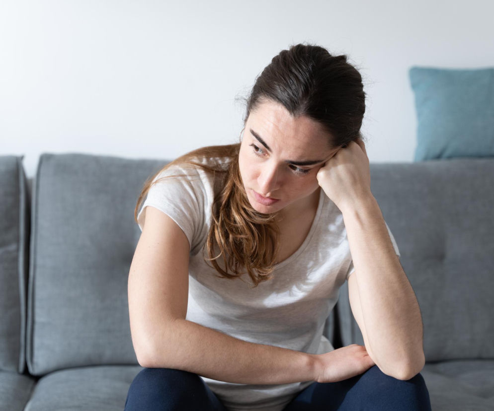 Zmęczona kobieta o długich, brązowych włosach siedzi na szarej kanapie w jasnym pomieszczeniu, z ręką pod brodą i smutnym wyrazem twarzy, odzwierciedlającym tematykę artykułu o mózgu głodowym. Dowiedz się więcej o hangry na Poradnik Zdrowie.