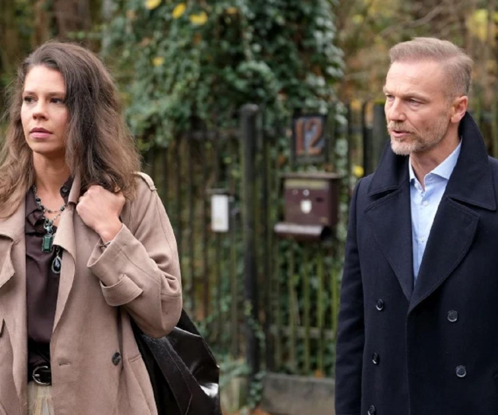 Maria Wieczorek jako Paulina Lipska w beżowym płaszczu i Andrzej Budzyński w granatowym płaszczu i jasnej koszuli stoją na zewnątrz, spoglądając w bok. Paulina, była ukochana Budzyńskiego, wraca, sprowadzając kłopoty z mężem gangsterem. Więcej na ten temat znajdziesz na Super Seriale.