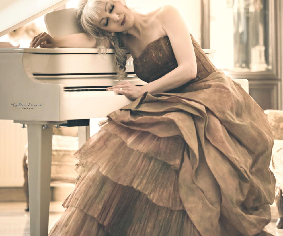 Pianistka Agnieszka Schulz-Brzyska w sukni wieczorowej opiera głowę o biały fortepian. O koncertach w USA pisze SE USA.