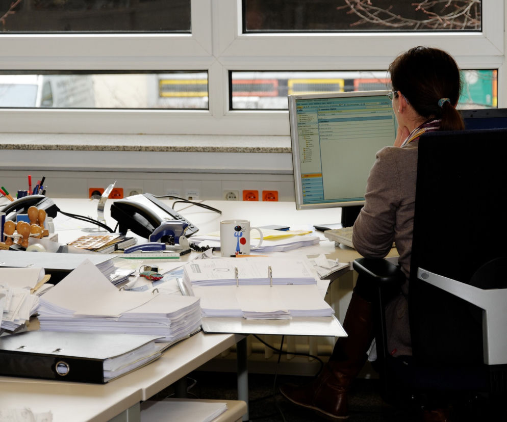 Kobieta w okularach pracuje przy komputerze w biurze pełnym dokumentów i sprzętu, symbolizującym biurokrację i zamieszanie wokół nowych przepisów o stażu pracy. Więcej o zmianach w Super Biznes.