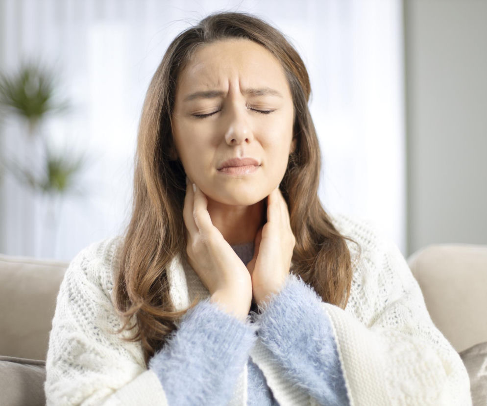 Młoda kobieta z zamkniętymi oczami dotyka dłońmi bolącego gardła, siedząc na kanapie w domu. Jej mimika twarzy wyraża dyskomfort i cierpienie. To typowa scena ilustrująca ból gardła, na temat którego można przeczytać na Poradnik Zdrowie.